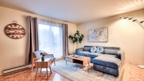A living room with a wooden floor, a blue sofa, a coffee table, a chair, a wall clock, and a window with curtains. - Montreal`s West Island Retreat! Value & Adventure! (Montréal)
