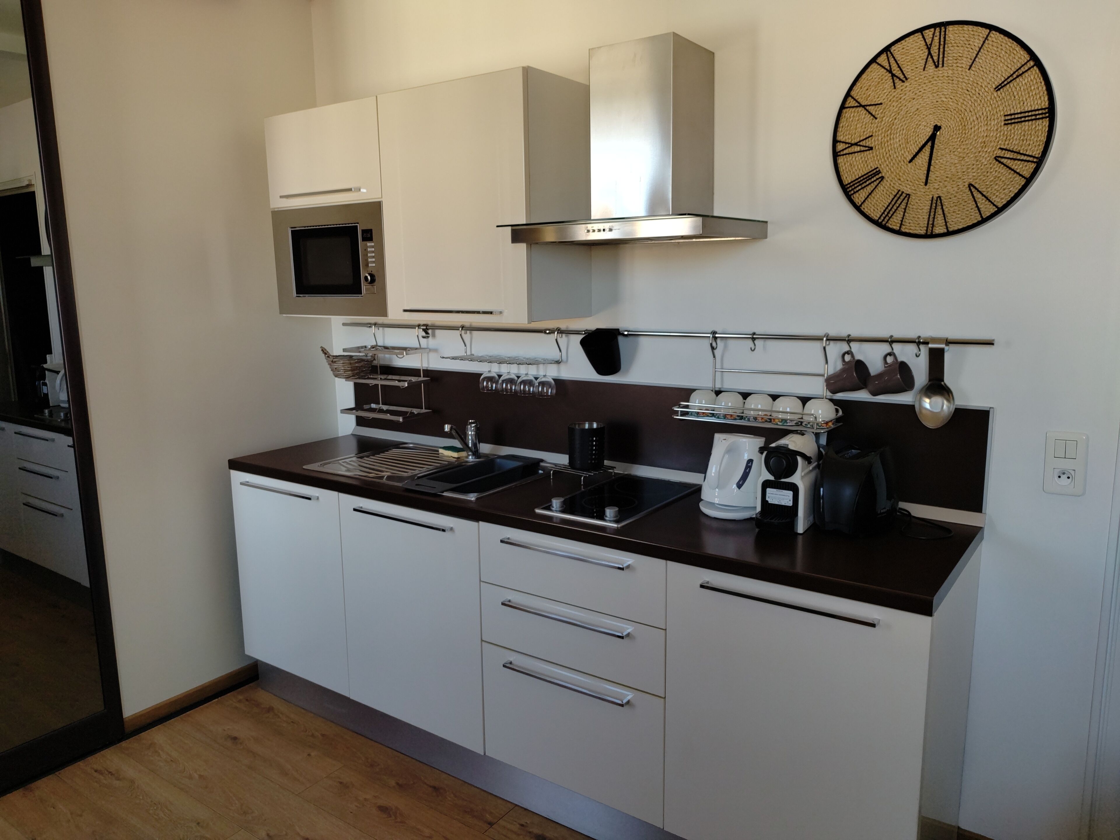 A modern kitchen with white cabinets, a stainless steel range hood, a microwave, a coffee maker, and a clock on the wall.