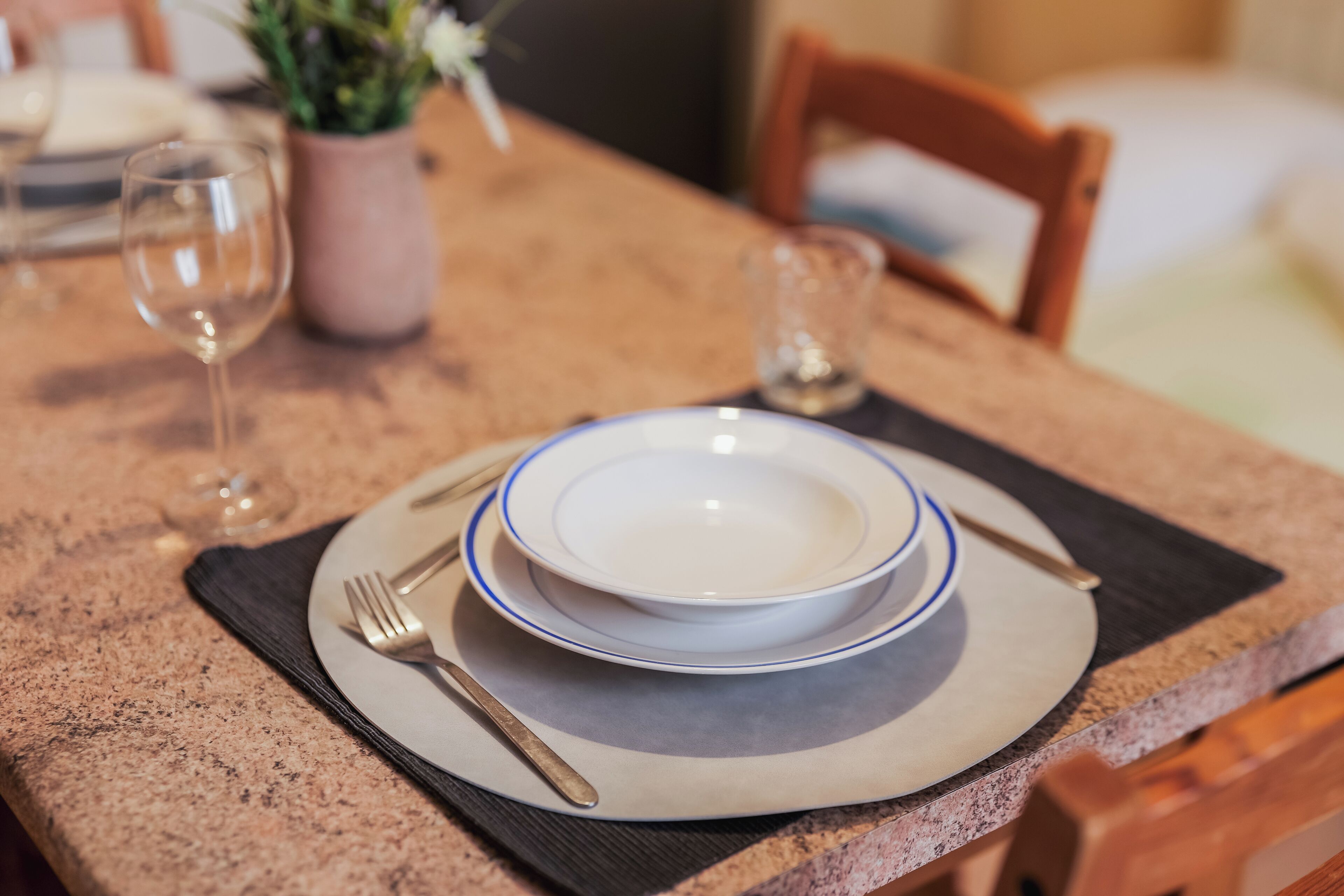 Una mesa de comedor dispuesta con platos, cubiertos y vasos, con un jarrón de flores al fondo.