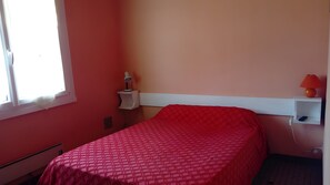 Une chambre avec un lit simple, une table de chevet avec une lampe, une étagère fixée au mur avec un téléphone, et une fenêtre avec un rideau.