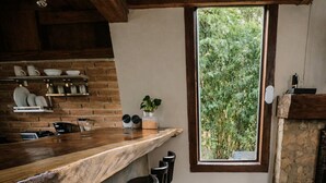 A bar area with wooden stools, a brick wall, and a view of greenery through a window.