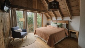 A cozy bedroom with a large bed, a chair, a TV, and a view of greenery outside.