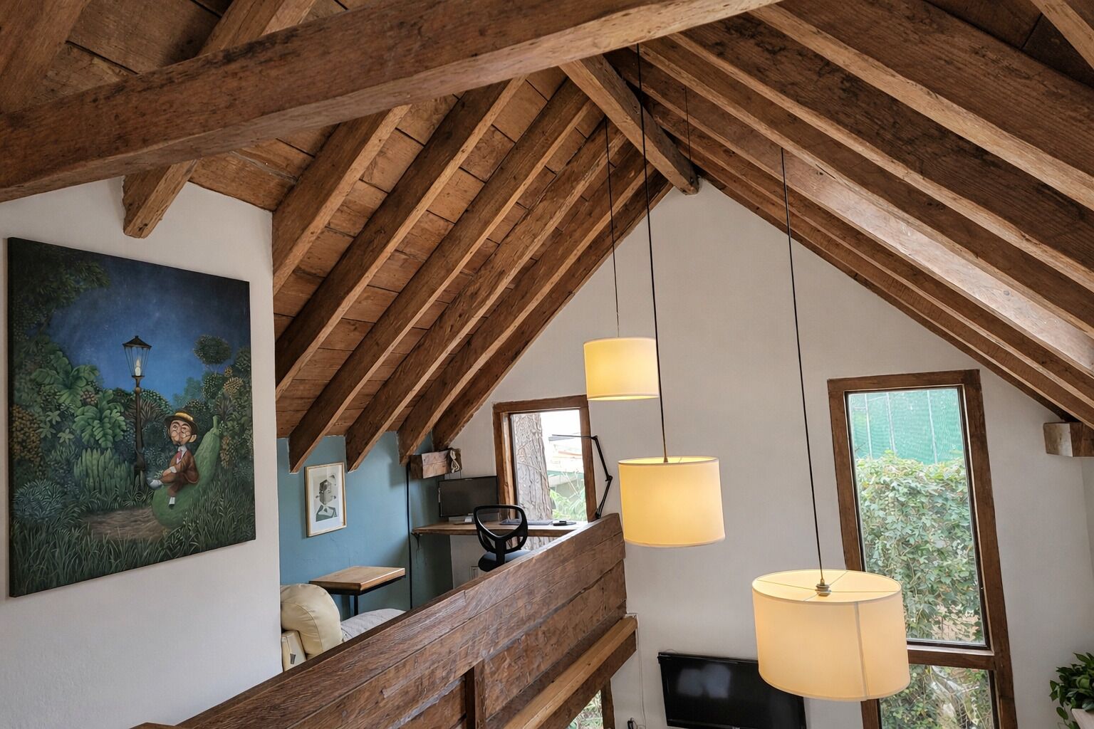A rustic attic room with exposed wooden beams, a desk with a computer, and a large window with a view of greenery.
