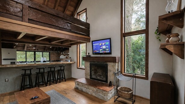 A rustic kitchen with a stone fireplace, wooden beams, and a large window overlooking greenery.