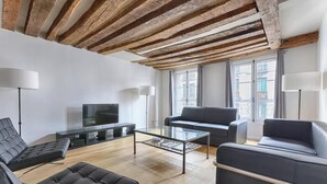 A modern living room with a wooden ceiling, a flat-screen TV, a glass coffee table, and two sofas. - PARISIAN ELEGANCEWITH EXPOSED BEAMS PERFECTLY LOCATED-SAINT GERMAIN– RUE DU BUCI (Paris)