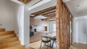 A modern kitchen with wooden beams, a dining area with a table and chairs, and a staircase leading to another level. - PARISIAN ELEGANCEWITH EXPOSED BEAMS PERFECTLY LOCATED-SAINT GERMAIN– RUE DU BUCI (Paris)