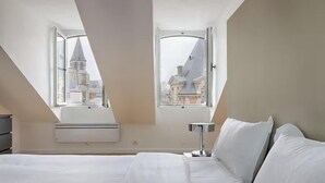 A hotel room with a bed, a bedside table, and two windows offering a view of buildings. - PARISIAN ELEGANCEWITH EXPOSED BEAMS PERFECTLY LOCATED-SAINT GERMAIN– RUE DU BUCI (Paris)