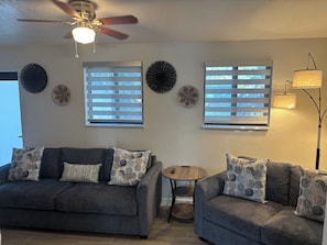 Ein Wohnzimmer mit zwei grauen Sofas, einem Deckenventilator und zwei mit Jalousien versehenen Fenstern.