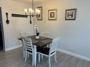 Un coin repas avec une table en bois foncé, des chaises blanches, un vase avec des fleurs blanches, un luminaire fixé au mur et des tableaux encadrés.
