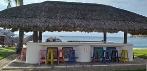 Eine Strandbar mit einem Reetdach, bunten Hockern und Blick auf das Meer.