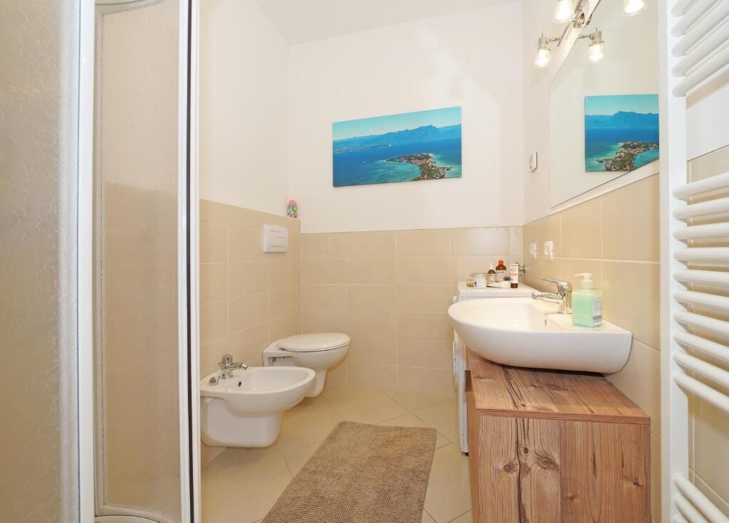 A bathroom with a shower, bidet, toilet, and sink. There are two wall-mounted pictures depicting a coastal landscape.