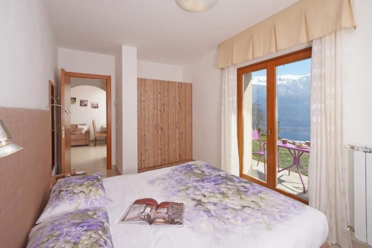 A bedroom with a bed, a wooden wardrobe, a bathroom visible through an open door, and a balcony with a table and chairs.