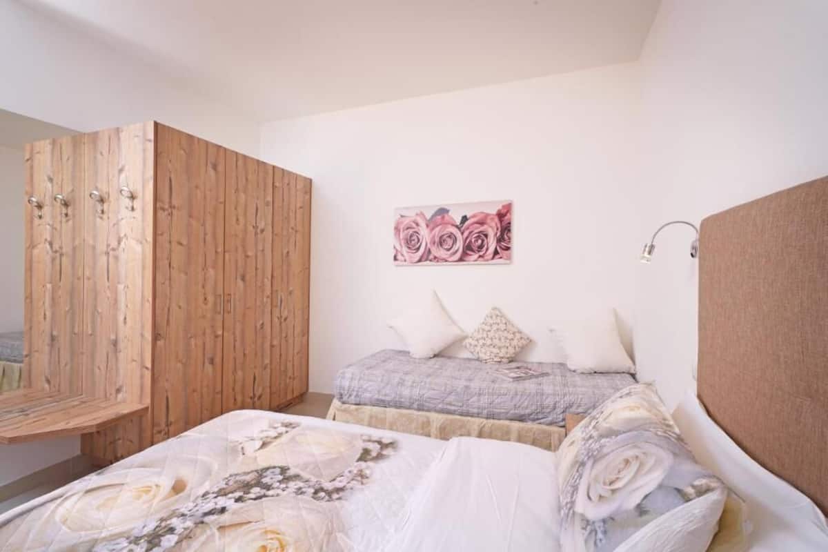 A bedroom with a wooden wardrobe, a bed with floral bedding, a grey quilted bedspread, and a pink rose painting on the wall.