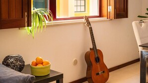 A room with a wooden door, a window with open shutters, a guitar, a small table with a bowl of oranges, and a plant. - Casa Da Bélgica: Sea View, WiFi & Air Conditioning in Sagres (Sagres)