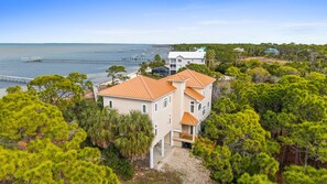 A two-story house with a red roof, surrounded by pine trees, near a body of water with a pier. - Canary On The Bay: Bayfront, Pool, Pet-friendly, Shared Dock, Sleeps 14 (St. George Island)