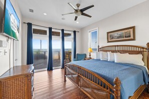 A bedroom with a large bed, a ceiling fan, a TV, and a view of the ocean through the sliding glass door. - Canary On The Bay: Bayfront, Pool, Pet-friendly, Shared Dock, Sleeps 14 (St. George Island)