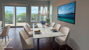 Un coin repas avec une table blanche, des chaises beiges et une grande fenêtre donnant sur l’extérieur.