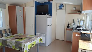A compact kitchen with a built-in TV, a dining area with a table, and a small seating area. - Mobile Home '4 Personnes Climatisé À' with Shared Pool, Private Terrace and Air Conditioning (Saint-Pierre-d'Oléron)