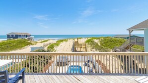 Blick auf einen Strand von einer Holzterrasse aus, mit Pool, Rettungsschwimmerstand und einem Haus im Hintergrund.