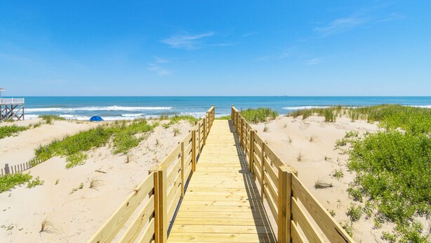 Ein hölzerner Steg führt zu einem Strand mit Dünen und wolkenlosem, blauem Himmel.