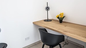 Ein moderner Arbeitsplatz mit einem Holztisch, einem schwarzen Stuhl, einer Stehlampe und einem Blumentopf mit gelben Blüten.
