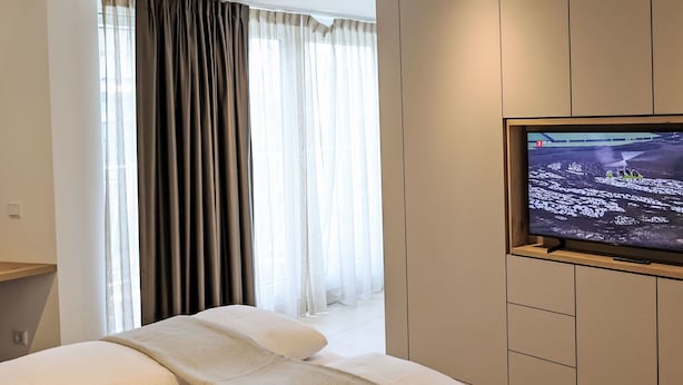 Ein modernes Hotelzimmer mit einem großen Flachbildfernseher an einem Holzschrank, einem Bett mit weißer Bettwäsche und einem großen Fenster mit transparenten Vorhängen.