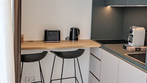 Eine kleine Küche mit Toaster, Kaffeemaschine und einem weißen Kaffeeautomaten auf der Arbeitsplatte. Darunter befinden sich zwei schwarze Barhocker.