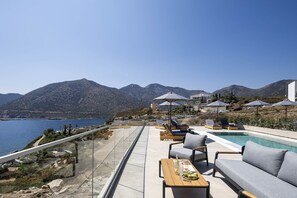 En tagterrasse med pool, liggestole og spisebord med udsigt over en bjergrig kystlinje.