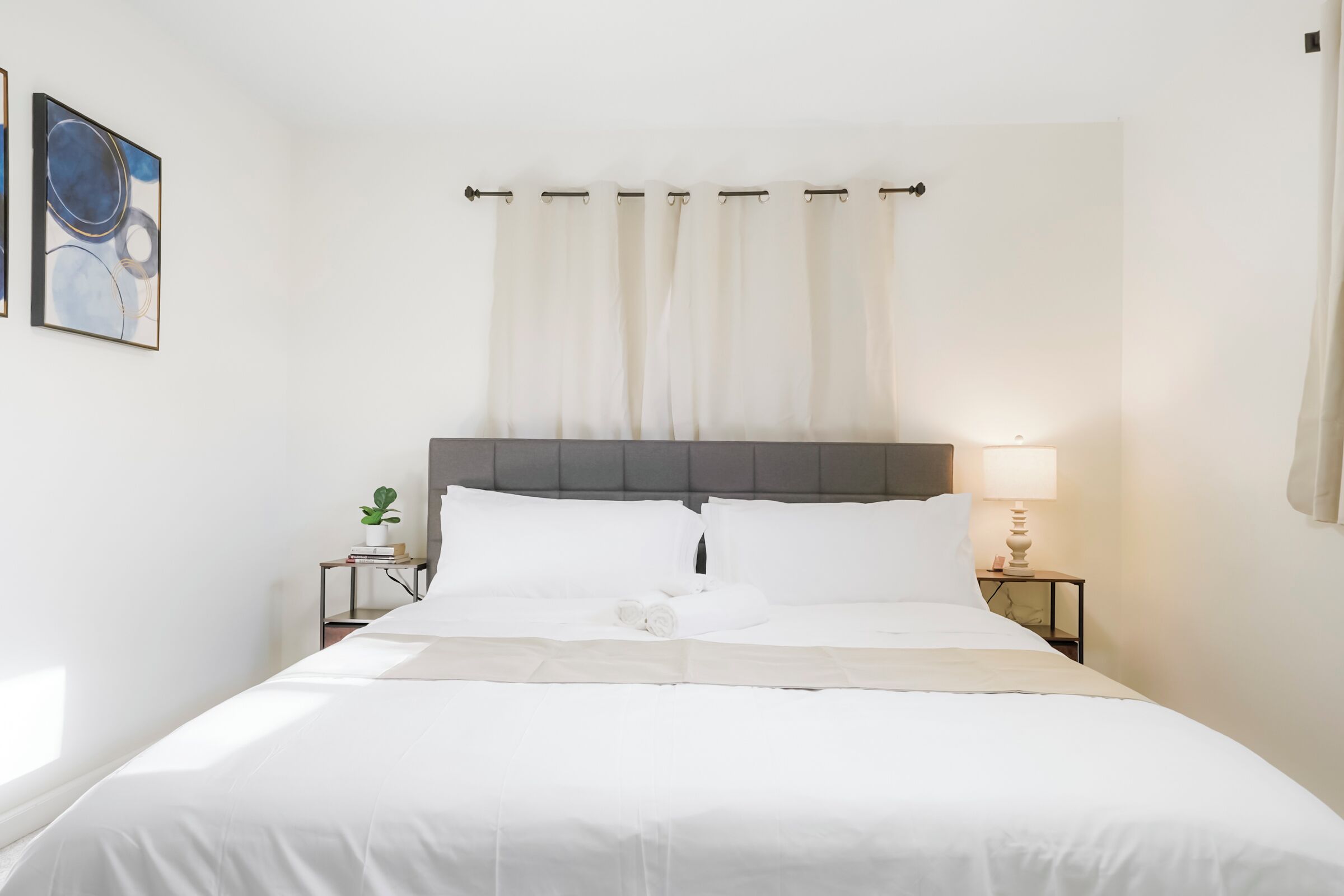 En pænt redt seng med hvide sengetæpper, en grå sengegavl og et natbord med en lampe.