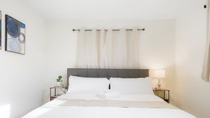 Een keurig opgemaakt bed met witte lakens, een grijs hoofdeinde en een nachtkastje met een lamp.