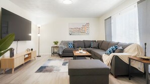 Een moderne woonkamer met een grijze bank, een flatscreen televisie aan de wand, een bijzettafeltje en een grote plant.