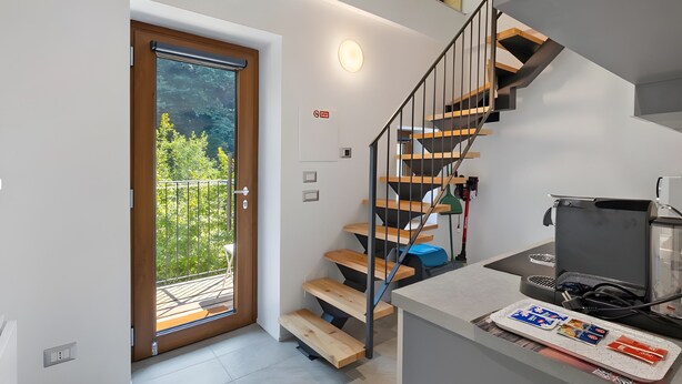 Ein modernes Interieur mit einer Holztreppe, einer kleinen Küchenzeile und einer Tür zu einem Balkon mit Blick auf Bäume.