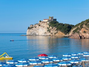 A coastal scene with a historic building on a cliff, calm sea, and beach umbrellas. - Prestige Family by Interhome (Castiglione della Pescaia)