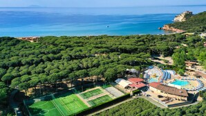 Vista aérea de un complejo turístico frente al mar, con cancha de tenis, piscina y abundante vegetación.