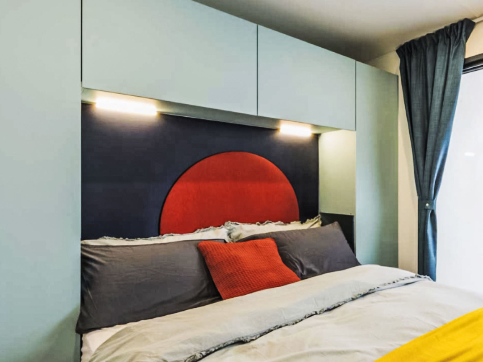 Ein modernes Schlafzimmer mit individuellem Kopfteil, einem Bett mit weißer und grauer Bettdecke und einem Fenster mit dunklen Vorhängen.