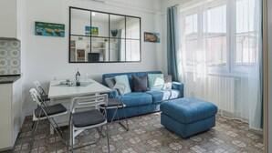Ein Wohnzimmer mit einem blauen Sofa, einem Esstisch mit Stühlen, einem großen Spiegel und einem Fenster mit Vorhängen.
