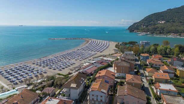 Eine Küstenstadt mit Strand, der mit Sonnenschirmen gesäumt ist, einer Marina und Wohngebäuden mit roten Ziegeldächern.