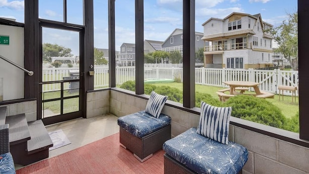 A covered outdoor seating area with wicker furniture and cushions, a view of a residential neighborhood, and a white picket fence. - 4BR Home Near the Beach! | Canal Access + Porch (South Bethany)