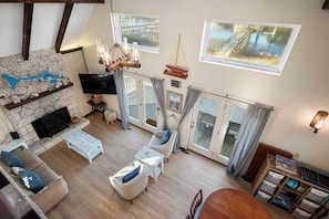 A spacious living area with a stone fireplace, comfortable seating, a television, and a view of a dock and water through large windows. - Intracoastal Escape: Coastal Cottage with Private Dock (Oak Island)