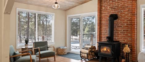 A cozy living room with a fireplace, wooden flooring, and large windows.