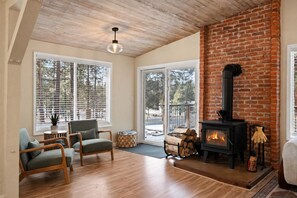 A cozy living room with a fireplace, wooden flooring, and large windows. - Private Sauna + Hot Tub - Mins to Mt. Bachelor (Bend)