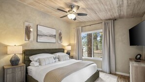 Un dormitorio con una cama, dos mesitas de noche, un ventilador de techo, una ventana con cortinas y un televisor en la pared.