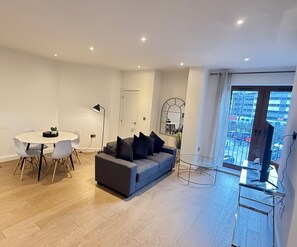 Een moderne woonkamer met een grijze bank, een ronde eettafel met stoelen, een kleine potplant en een glazen salontafel.