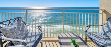 A balcony with two chairs and a table, overlooking a beach with waves and a clear sky.