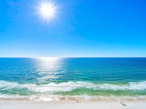 A beach with clear blue water, a sandy shore, and a bright sun in the sky. - Emerald Beach 1831 (Panama City Beach)