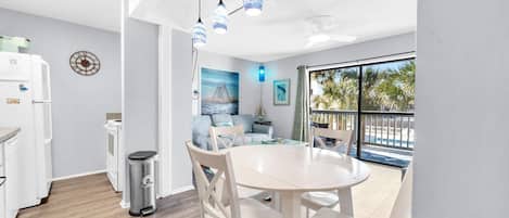 A modern kitchen with white appliances, a dining area with a round table and chairs, and a large window with a view of palm trees.