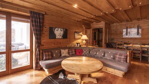 A cozy cabin interior with a wooden ceiling, a large window with curtains, a sofa with plaid cushions, a round wooden table, and a painting on the wall. - Casa Merisanda, comfortable and surrounded by Natur (Cortina d´Ampezzo)