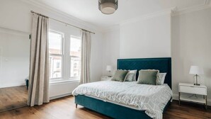 Makuuhuoneessa on turkoosinsininen sänky, valkoiset vuodevaatteet, kaksi yöpöytää, joissa on lamput, ja ikkuna, jossa on verhot.