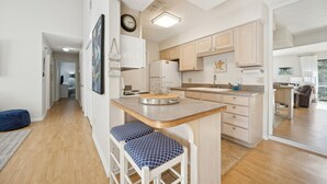 A modern kitchen with wooden cabinets, a built-in island with bar stools, a refrigerator, and a clock on the wall. - Amelia Island 2 Bedroom Getaway located in Forest Ridge (Fernandina Beach)