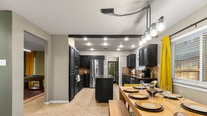 A modern kitchen with dark cabinets, a central island, and a dining area with a wooden table set for four. - Ultimate Getaway • Pool • Game Room • Sleeps 12 (San Antonio)